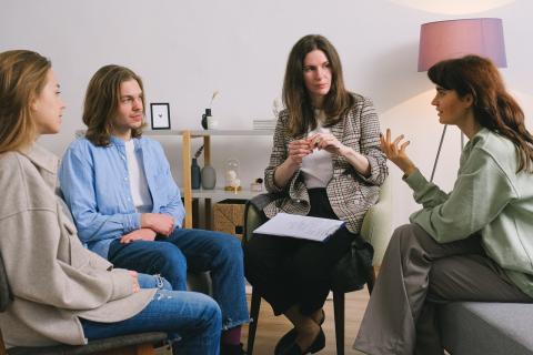 A group of people in discussion with a doctor, who has paper ready to make notes. The doctor is relatively casually dressed, and there is no desk between the doctor and the others.