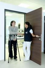 A medical professional talking with a patient as they walk through a doorway on crutches