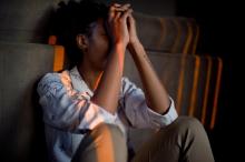 A woman sits on the ground against a wall, knees up to her chest and hands in front of her face.